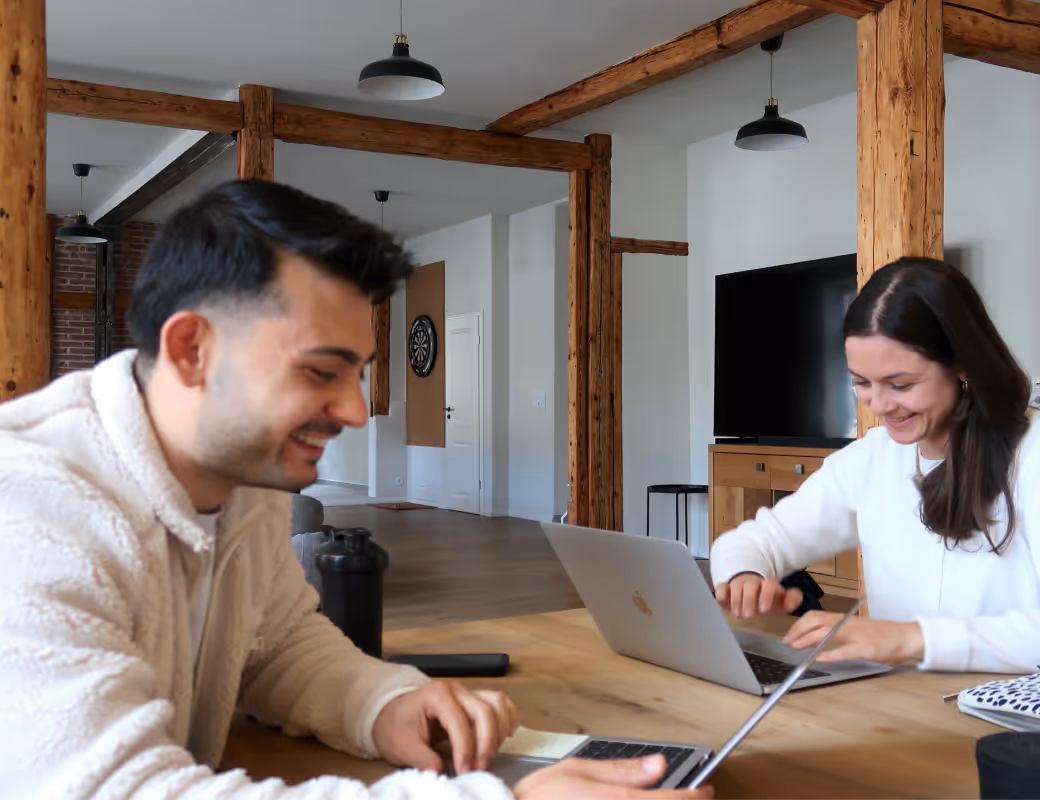Zwei Personen lächeln und arbeiten an Laptops an einem Holztisch in einem Raum mit sichtbaren Holzbalken und einer Dartscheibe an der Wand.