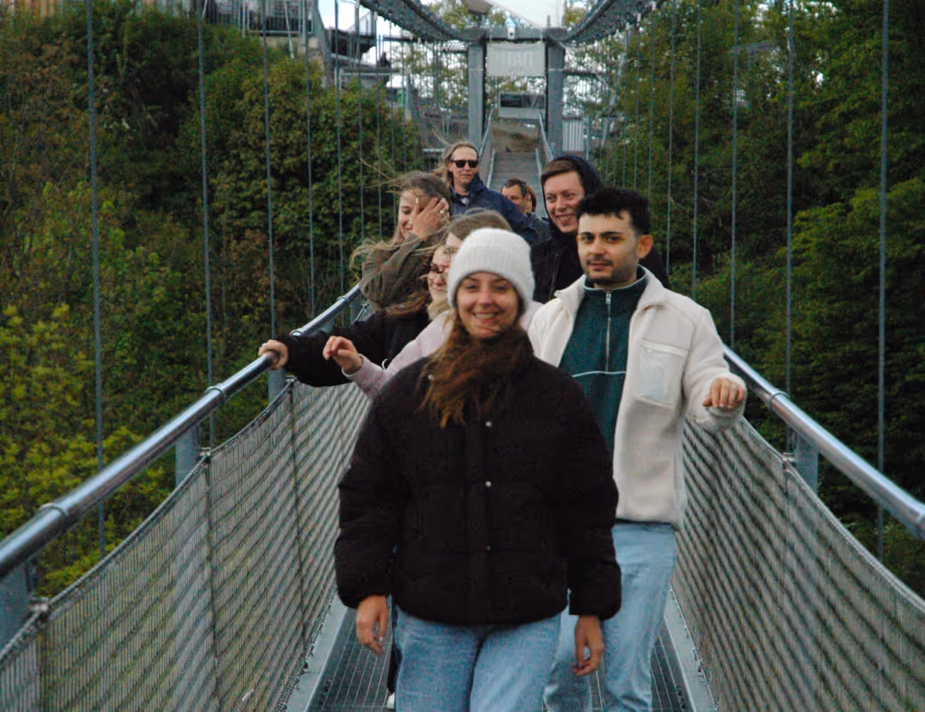 Gruppe von Menschen, die über eine Hängebrücke gehen, umgeben von grünen Bäumen, wobei eine Frau mit weißer Mütze vorneweg geht.