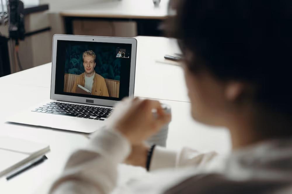 Person sitzt an einem Tisch und nimmt an einem Videoanruf auf dem Laptop teil, auf dem ein Mann mit Notizblock zu sehen ist.