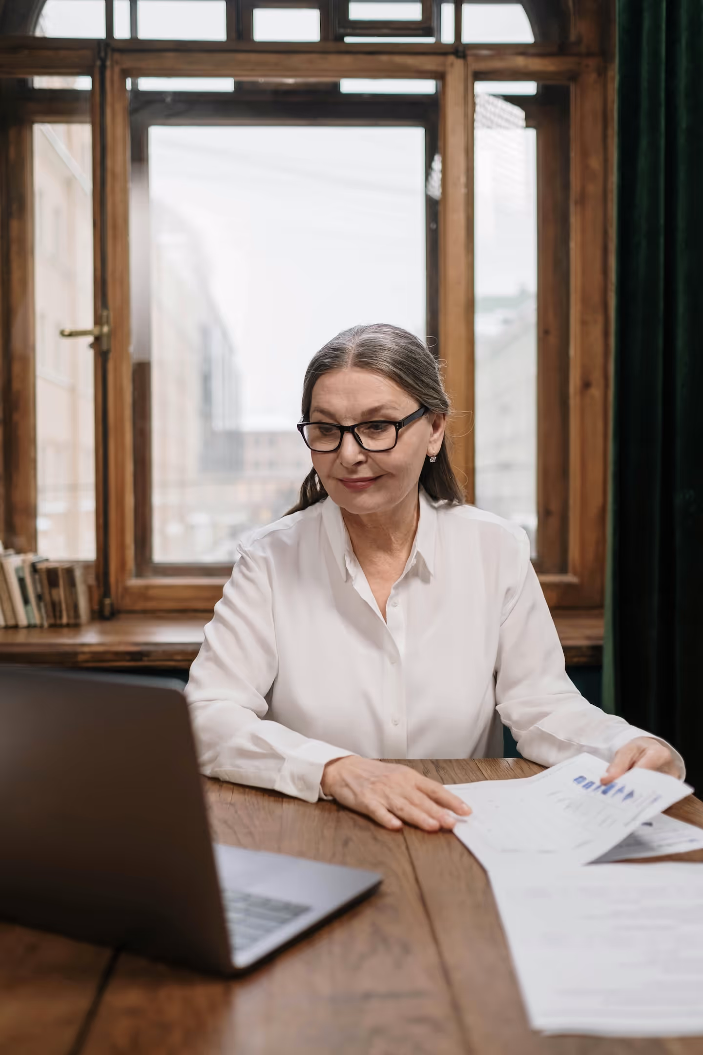 Ältere Frau mit Brille hält Papiere mit Diagrammen in der Hand und sitzt an einem Laptop.