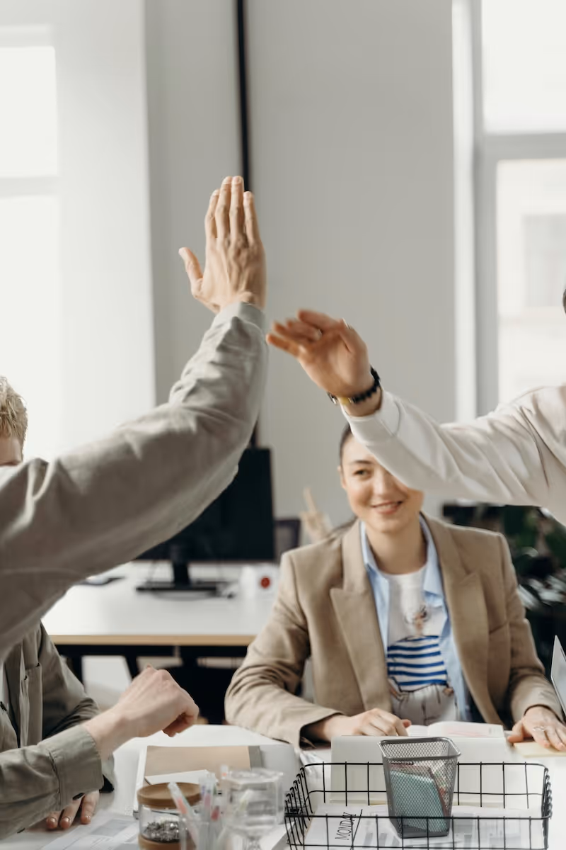 Zwei Personen geben sich ein High-Five über einem Arbeitstisch, während eine dritte Person im Hintergrund lächelt.