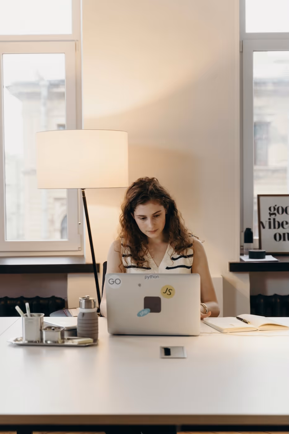Frau mit lockigem Haar arbeitet konzentriert an einem Laptop mit Entwickler-Stickern, an einem Schreibtisch mit Notizbuch und Wasserflasche.