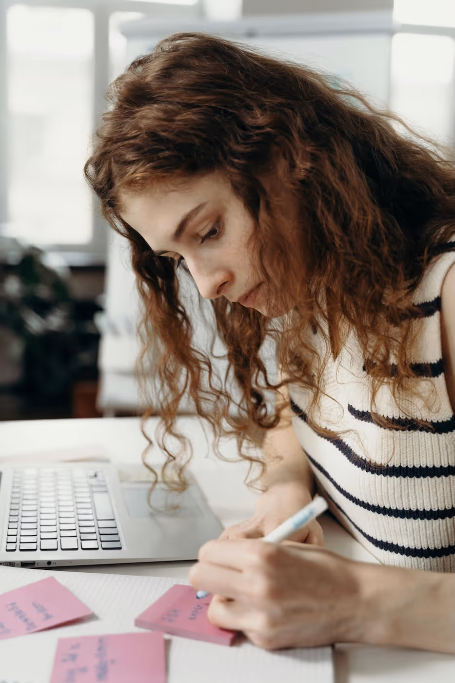 Junge Frau mit lockigem Haar schreibt mit einem Stift auf ein pinkes Haftnotiz-Zettelchen, ein Laptop steht daneben auf dem Tisch.
